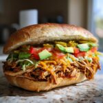 A close-up of a fully loaded Chicken Torta Sandwich featuring shredded chicken, cheese, avocado, tomatoes, and jalape&ntilde;os.