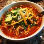 Close-up of a bowl of vibrant red Chicken Tortilla Soup topped with shredded chicken, avocado, cheese, and tortilla strips.
