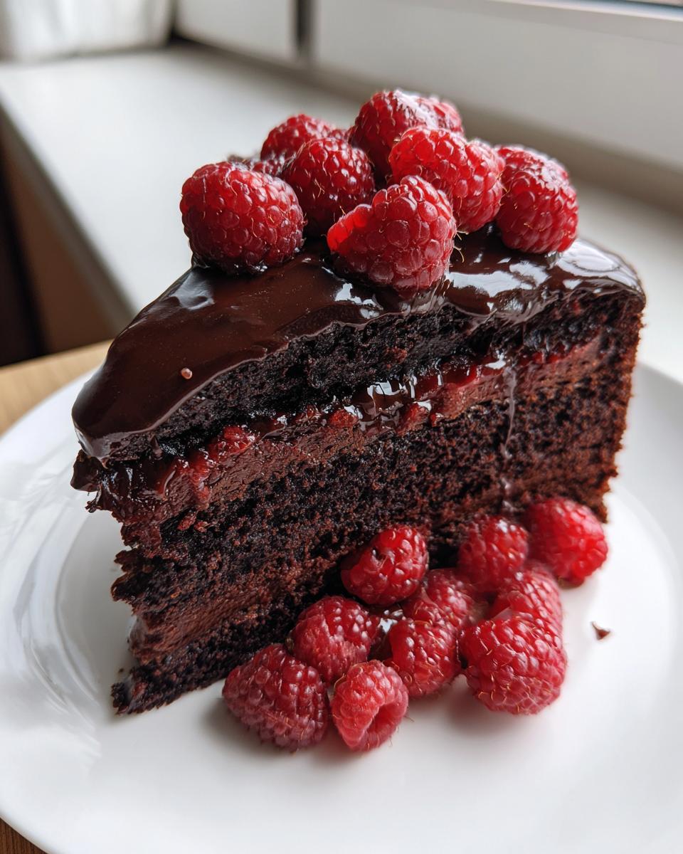 A rich slice of Chocolate Cake With Chocolate Ganache And Raspberries, topped and garnished with fresh red raspberries.