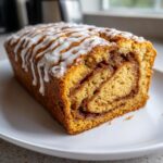 A close-up view of a freshly baked Cinnamon Roll Pumpkin Bread loaf with a visible swirl and white icing drizzle.