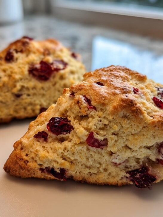 Two freshly baked Cranberry Orange Scones, golden brown with visible dried cranberries, resting on a light plate.