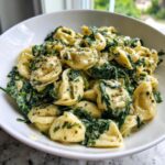 A close-up of creamy Garlic Spinach Tortellini tossed in a light sauce and seasoned with herbs, served in a white bowl.