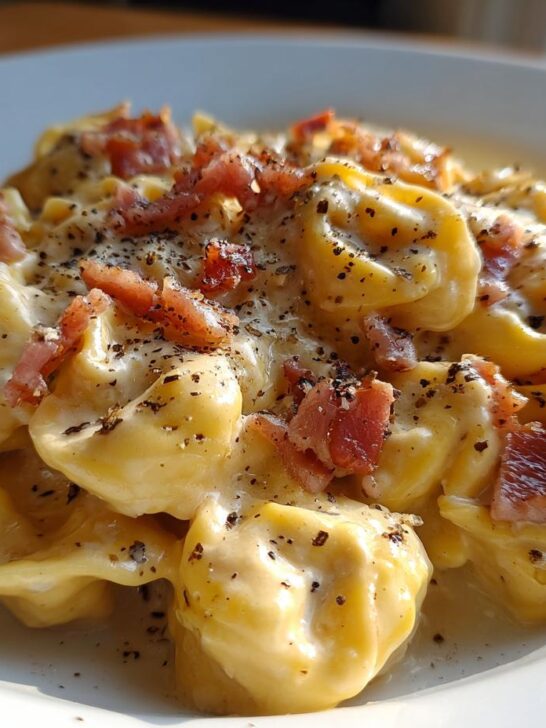 A close-up of creamy Tortellini Carbonara pasta topped with crispy bacon pieces and cracked black pepper.
