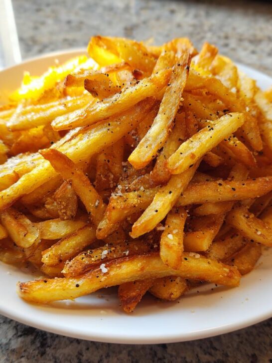A close-up, featured image of a pile of golden, crispy seasoned French fries sprinkled with salt and pepper.