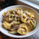 Close-up of a white bowl filled with creamy Crock Pot Cheesesteak Tortellini, featuring cheese, steak, and green peppers.