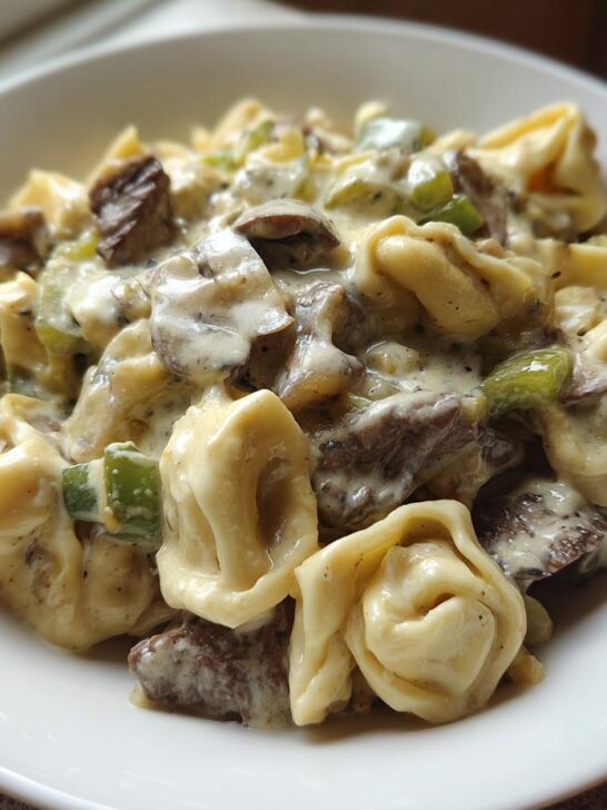A close-up of a white bowl filled with creamy Crock Pot Cheesesteak Tortellini, featuring beef, peppers, and mushrooms.
