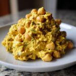 A close-up of a generous mound of bright yellow Curried Chickpea Salad topped with whole chickpeas, served on a white plate.