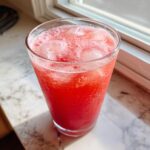 A tall glass filled with bright red Easy Homemade Strawberry Lemonade and ice cubes, sitting on a sunny countertop.