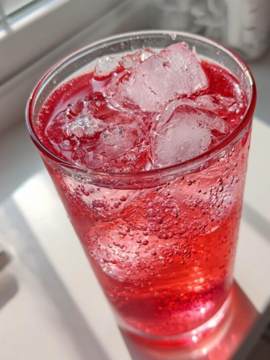 Close-up of a tall glass filled with bright red, fizzy Easy Homemade Strawberry Lemonade and ice cubes.