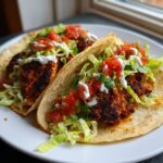 Two fully loaded Easy Weeknight Blackened Chicken Tacos on a white plate, topped with shredded lettuce, salsa, and crema.