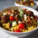 Close-up of a vibrant Greek Chicken Bowls featuring seasoned chicken pieces over yellow quinoa, topped with cherry tomatoes, cucumber, feta, and olives.