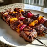 Close-up of two perfectly grilled steak kabobs featuring charred beef cubes, red onion, and yellow bell peppers.