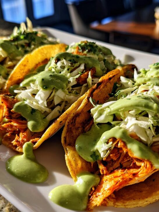 Close-up of three Jerk Salmon Tacos topped with slaw and drizzled generously with avocado cilantro lime sauce.
