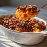 A spoonful of bright orange Maple Pecan Sweet Potato Casserole lifted above the white baking dish.