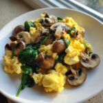 Fluffy yellow scrambled eggs mixed with saut&eacute;ed mushrooms and wilted spinach, served on a white plate.