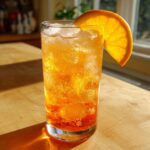 A tall glass filled with an effervescent Orange Crush Mocktail, ice cubes, and garnished with a fresh orange slice.
