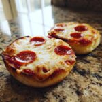 Two freshly baked pepperoni English Muffin Pizzas with melted cheese sitting on a granite countertop.