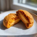 A Pumpkin Cheesecake Cookie broken in half showing the thick, creamy filling inside the soft pumpkin cookie.