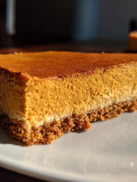Close-up of a thick slice of Pumpkin Spice Cheesecake Bars showing the graham cracker crust and creamy orange filling dusted with spice.