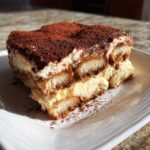 A rich slice of Pumpkin Spice Tiramisu showing layers of cream and coffee-soaked ladyfingers, topped with cocoa powder.