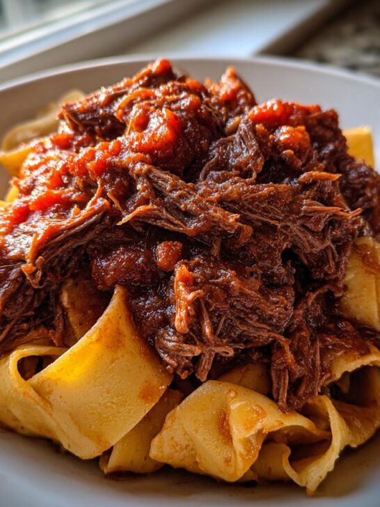 A close-up of tender, shredded Slow Cooker Beef Ragu piled high on wide pappardelle pasta in a white bowl.