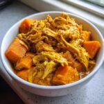 Close-up of shredded chicken and large chunks of sweet potato in a rich yellow sauce, served in a white bowl, featuring Slow Cooker Chicken And Sweet Potato Curry.