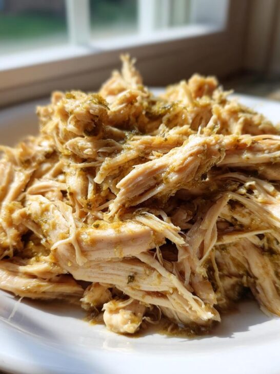 A close-up of shredded Slow Cooker Salsa Verde Chicken piled high on a white plate near a sunny window.