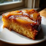 A close-up of a moist slice of Spiced Pear Upside Down Cake on a white plate, featuring caramelized pear slices on top.