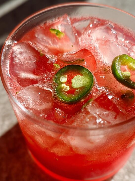 Close-up of a vibrant red Spicy Watermelon Mojito Mocktail filled with ice and garnished with two slices of green jalape&ntilde;o.