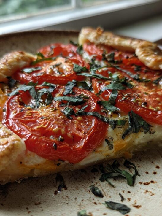 Close-up of a slice of Tomato Basil Galette featuring roasted red tomatoes over cheese on a flaky crust, garnished with fresh basil.
