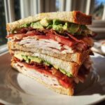 A tall, stacked Turkey Avocado Club Sandwich cut in half, showing layers of turkey, bacon, avocado, and tomato on toasted bread.
