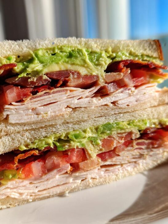 A cross-section view of a tall Turkey Avocado Club Sandwich stacked high with turkey, bacon, tomato, and mashed avocado on white bread.