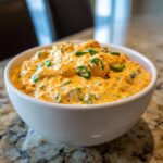 A close-up of a white bowl filled with creamy, orange Vegan Jalapeno Queso Dip, topped with sliced green jalapenos.