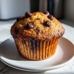 A single, perfectly baked Zucchini Chocolate Chip Muffin topped with melted chocolate chips on a small white plate.