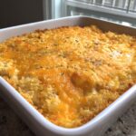 A close-up of a freshly Baked Cheesy Chicken Casserole topped with melted orange cheddar and dried herbs in a white square baking dish.