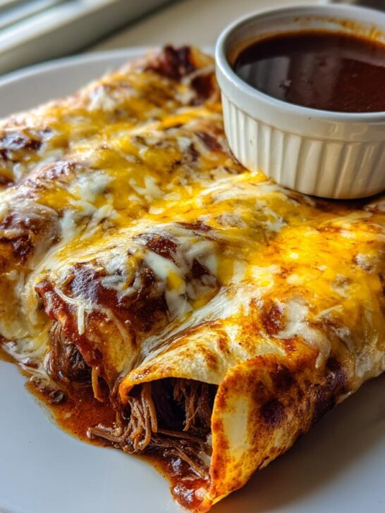 Two cheesy Birria Enchiladas covered in sauce, served with a side of consomm&eacute; for dipping.