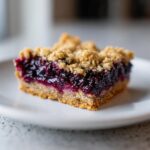 A close-up shot of a single, thick Blueberry Oatmeal Bars slice showing the crumbly oat topping and rich, dark blueberry filling.