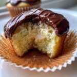 Close-up of a Boston Cream Pie Cupcakes with a bite taken out, revealing vanilla cream filling and chocolate ganache topping.
