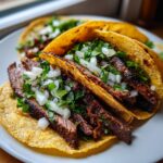 Close-up of two grilled Carne Asada Tacos filled with sliced marinated steak, topped generously with diced white onion and cilantro.