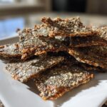 A close-up, sunlit stack of homemade, crispy Chia Flax Seed Crackers piled high on a rectangular white serving plate.