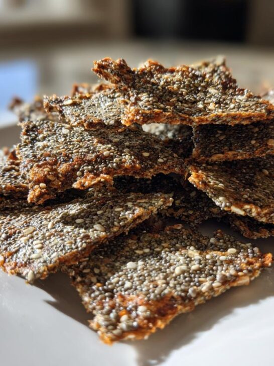 A close-up, sunlit stack of homemade, crispy Chia Flax Seed Crackers piled high on a rectangular white serving plate.
