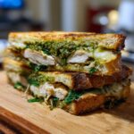 Two halves of a grilled Ciabatta Chicken Pesto Sandwich stacked on a wooden board, showing chicken, melted cheese, and green pesto.