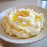A close-up of creamy Roasted Garlic Mashed Potatoes served in a white bowl with melted butter pooling on top.