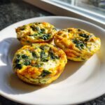 Three freshly baked Egg Muffins With Spinach And Feta resting on a white plate near a window.