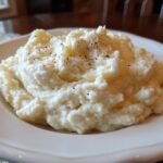 A generous serving of fluffy Mashed Cauliflower With Sour Cream, topped with cracked black pepper, on a white plate.