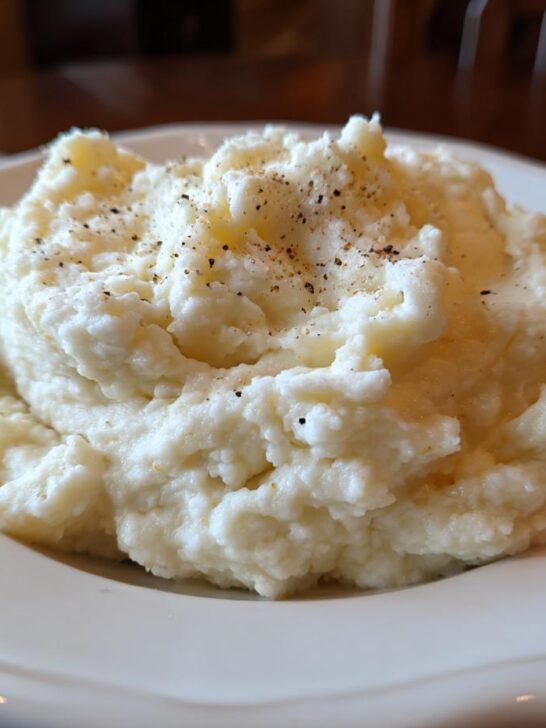 A generous serving of fluffy Mashed Cauliflower With Sour Cream, topped with cracked black pepper, on a white plate.