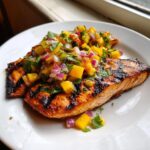 Two perfectly grilled salmon fillets topped generously with vibrant mango salsa, featuring diced mango, red onion, and cilantro.
