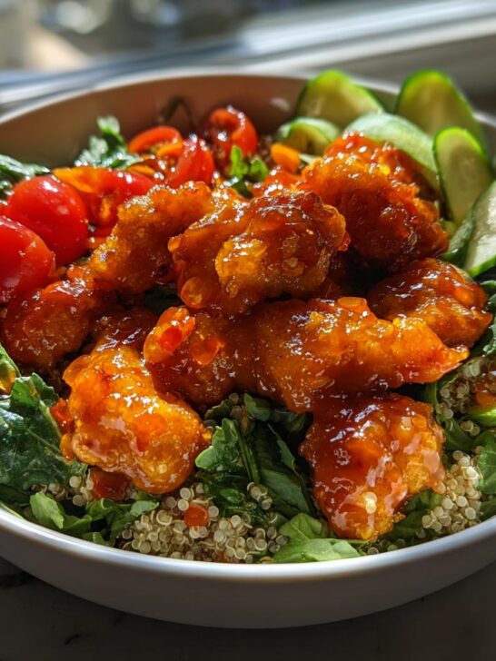 A bowl featuring crispy, glazed Hot Honey Chicken Bowl Sweet Green Copycat served over quinoa, greens, cucumbers, and cherry tomatoes.