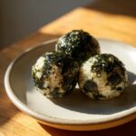 Three perfectly shaped Korean Rice Balls coated in crumbled seaweed and sesame seeds, resting on a light plate.