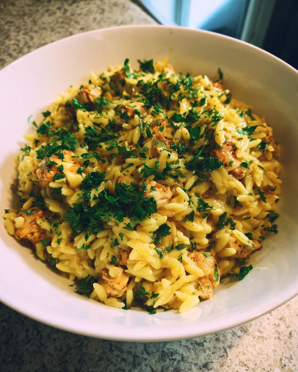 A close-up view of a white bowl filled with creamy Lemon Chicken Orzo, topped generously with fresh chopped parsley.